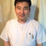 白い施術着を着て、ほほえむ松前雅義
