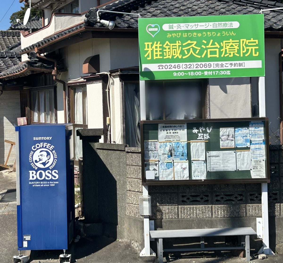 瓦屋根の建物の前に、緑色の看板「雅鍼灸治療院」が立っている。看板の下に掲示板とベンチ、左に青い自動販売機がある