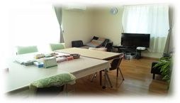 明るい部屋に白い長テーブルといすが並び、奥にテレビとソファがある
